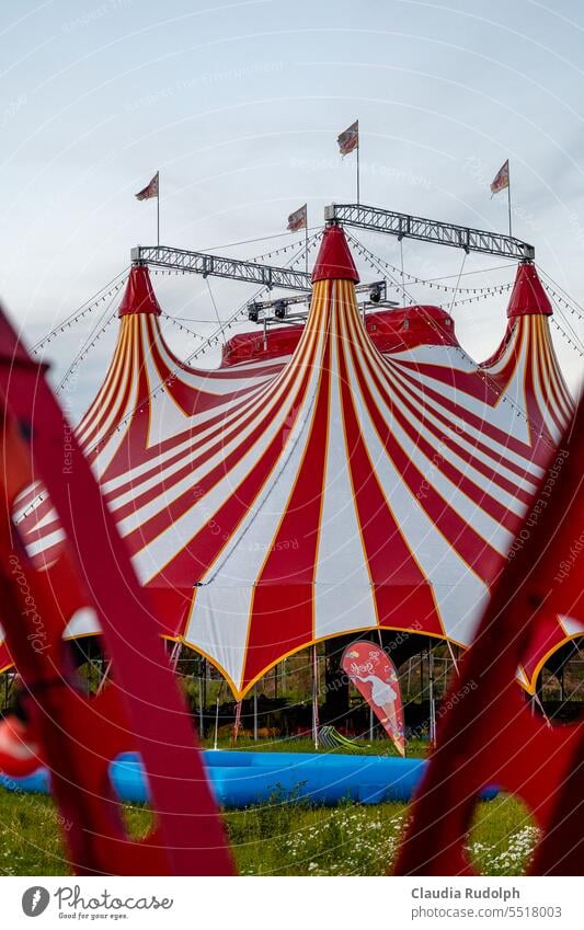 Blick durch Zaun auf ein rot weiß bestreiftes Zirkuszelt mit Lichterketten und Fahnenmasten Zirkus-Hintergrund Zirkuszelte Streifen Zelt Show Freizeit & Hobby