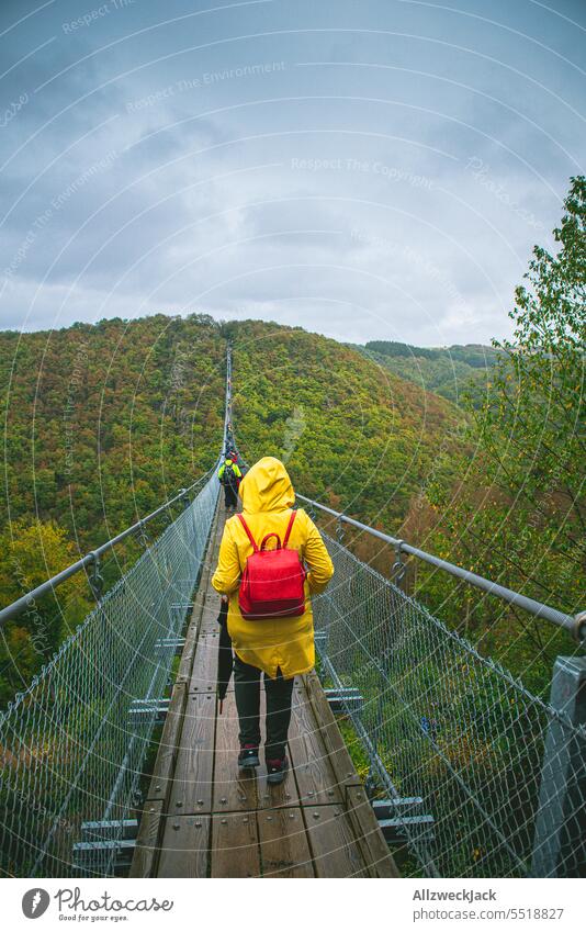 juneg Frau in gelbem Regenmantel läuft bei schlechtem Wetter über die Geierlay Hängebrücke schlechtes Wetter nass Regenjacke grau Brücke Ausflug