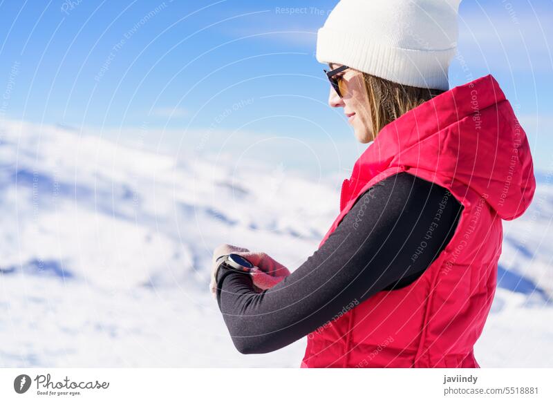 Junge Frau steht in verschneitem Gelände und prüft die Zeit Winter Armbanduhr prüfen Berghang Abenteuer warme Kleidung Schnee Berge u. Gebirge jung Tageslicht