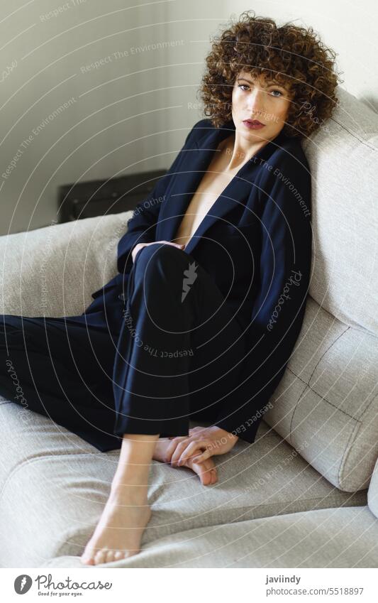 Stilvolle Frau mit lockigem Haar sitzt auf dem Sofa und trägt einen schwarzen Anzug selbstbewusst charismatisch Körperhaltung krause Haare Charme Outfit modern