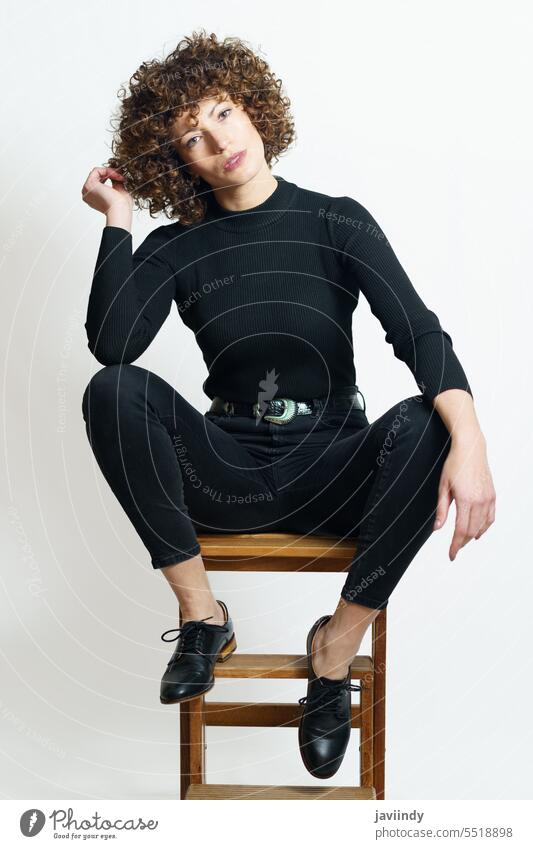 Stilvolle Frau in schwarzem Kleid sitzt auf einer Holzbank Hocker Haare berühren Mode hölzern Schritt Neigung Bein angehoben krause Haare sich[Akk] entspannen