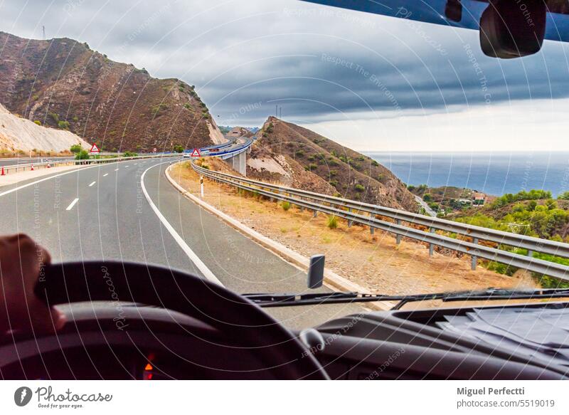 Blick aus der Fahrerposition eines Lastwagens auf eine Autobahn, die zwischen Bergen und dem Meer im Hintergrund verläuft. Innenbereich im Inneren