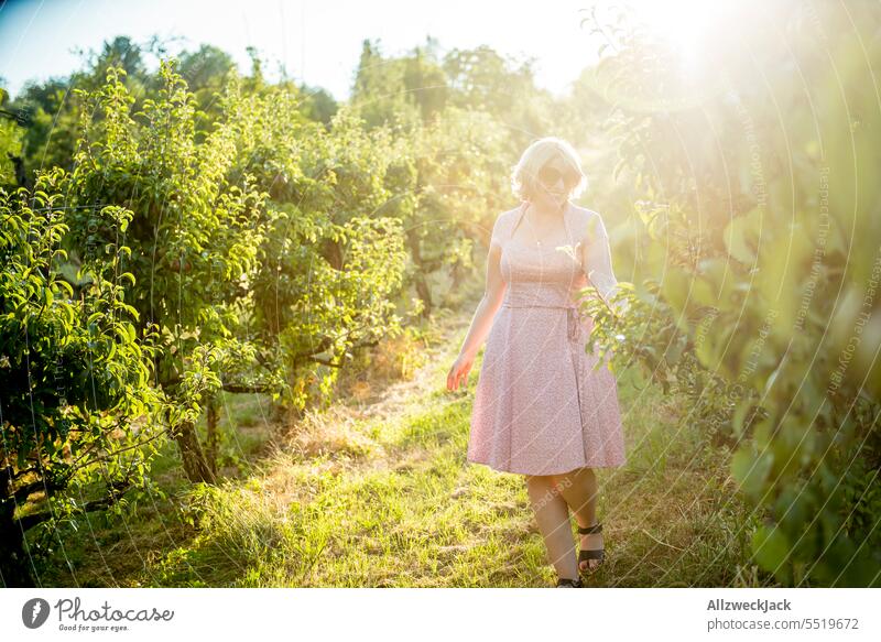 blonde Frau mittleren Alters mit rosa Kleid in einer Obstplantage zwischen Birnenbäumen schön hübsch Sympathie sympathisch Birnenbaum Sonnenlicht Sonnenstrahlen