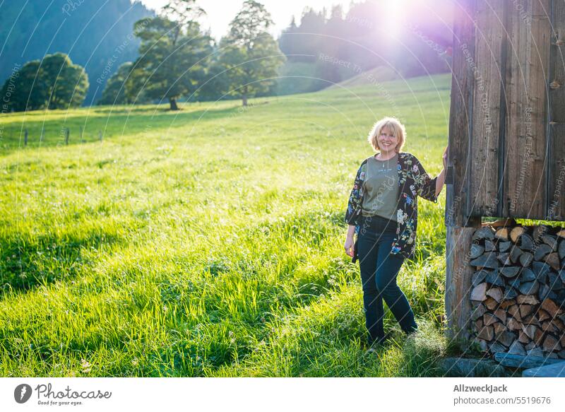 blonde Frau mittleren Alters sympathisch lächelnd im Gegenlicht an einem Chalet auf einer schweizer Alm Schweiz Schweizer Alpen Wiese Natur grün saftig