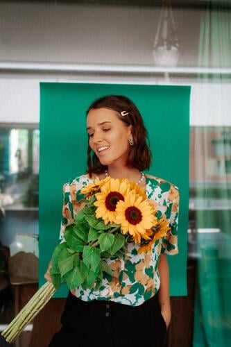 eine junge Frau mit einem Sonnenblumenstrauss in der Hand  und einem lächeln auf dem Gesicht. Sie trägt eine grün, gelbe Blouse und schwarze Hosen. Blumenstrauß