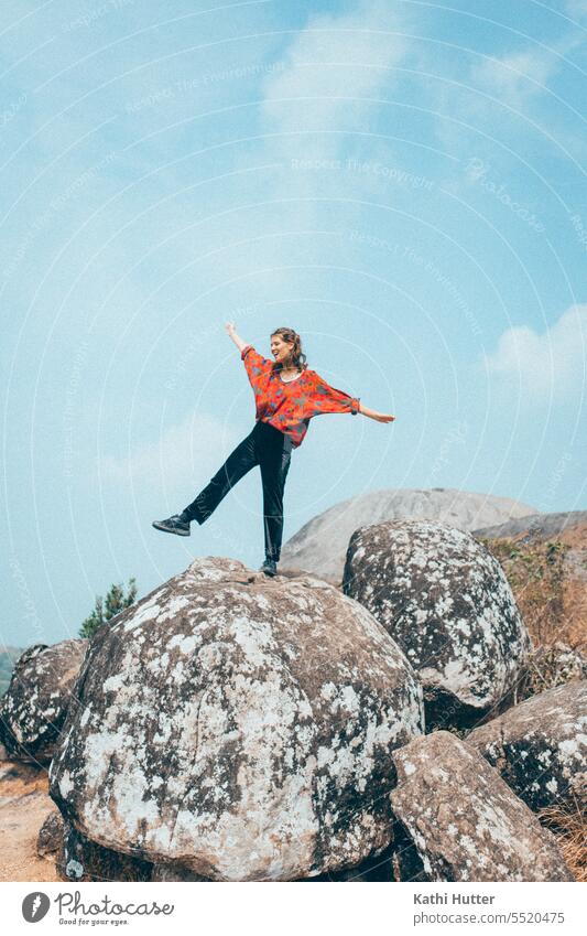 eine junge Frau steht auf einem grossen Stein, dahinter strahlt der blaue Himmel. Sie trägt eine rote Bluse und schwarze Hosen. Wolken Sommer Sonne