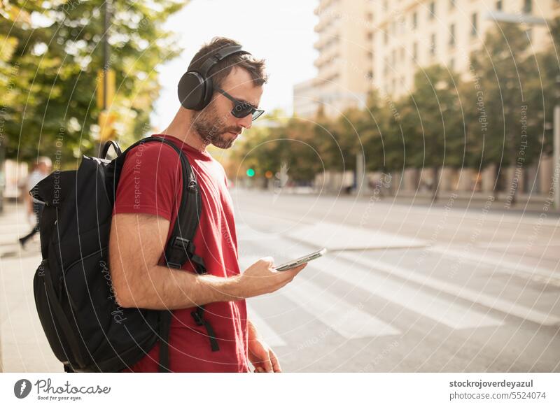 Ein junger Mann trägt eine Sonnenbrille, hört Musik und schaut auf sein Smartphone, während er eine Stadt in Spanien besucht. Erwachsener Lifestyle männlich