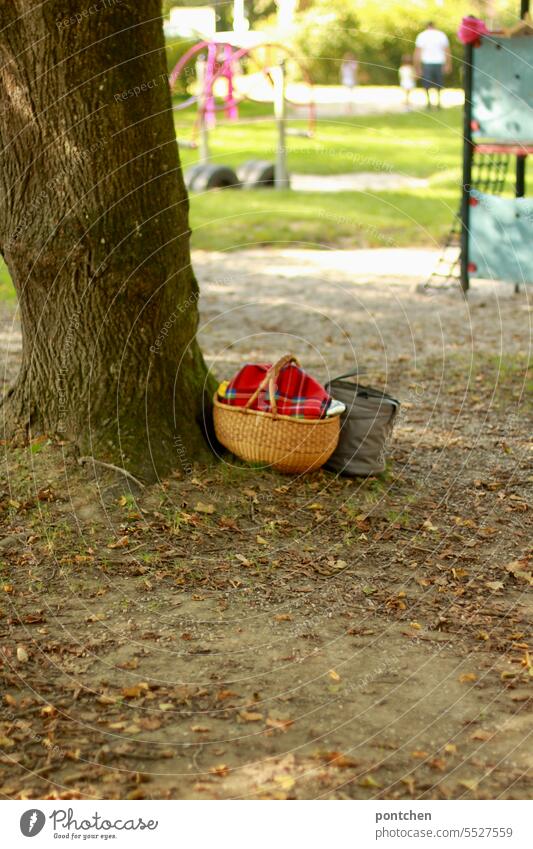 ein picknickkorb mit karierter decke steht an einem baum auf einem spielplatz . ein vater mit seinen beiden kindern im hintergrund proviant vorbereitet kindheit