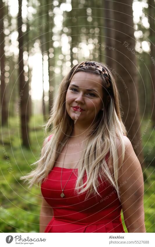 Portrait einer schönen, blonden, langhaarigen Frau, die in einem lichten Wald steht und in die Kamera schaut blonde Haare lange Haare rotes Kleid Pflanze grün