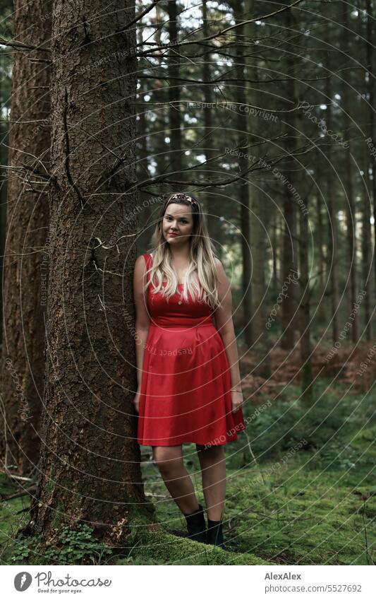 Portrait einer schönen, blonden, langhaarigen Frau, die in einem lichten Wald an einem Baum gelehnt steht und in die Kamera schaut blonde Haare lange Haare