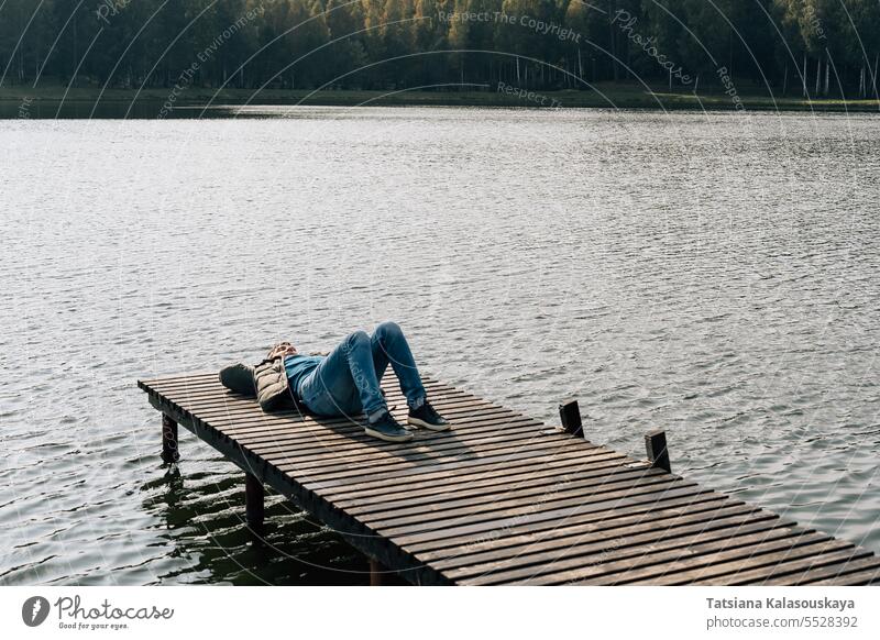 Ein Mann liegt entspannt auf einem hölzernen Bootssteg an einem See im Herbst Jeanshose Trägerkleid Jacke Lügen entspannend Bootsanlegestelle Dock genießend