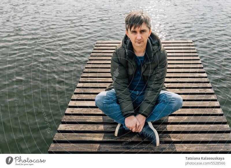 Ein erwachsener Mann sitzt in Jeans und Jacke im Schneidersitz auf einem Holzsteg an einem See Jeanshose Trägerkleid sitzen entspannend hölzern