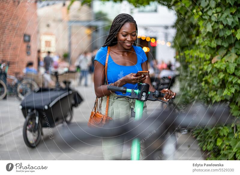 Porträt einer glücklichen jungen Frau, die ihr Smartphone benutzt, während sie mit einem Elektroroller in der Stadt steht Erwachsener attraktiv schön schwarz