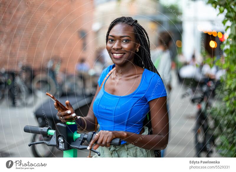 Porträt einer glücklichen jungen Frau, die ihr Smartphone benutzt, während sie mit einem Elektroroller in der Stadt steht Erwachsener attraktiv schön schwarz