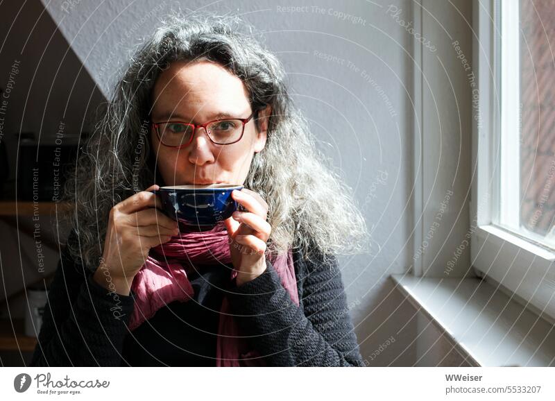 Eine Frau um die 50 sitzt im Licht am Fenster, trinkt Tee und fühlt sich wohl Schale Tasse trinken Getränk Heiß´getränk vergnügt zufrieden lächeln Blickkontakt