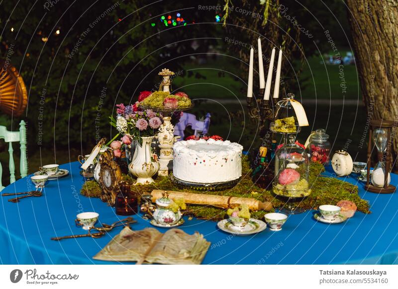 Der Esstisch ist im Stil von Alice im Wunderland dekoriert. Ein aufgeschlagenes altes Buch, ein Kuchen, Teeschalen, Kerzen in einem Kerzenständer, eine Sanduhr auf einer blauen Tischdecke spät am Abend