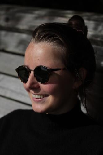 Schöne Frau mit Sonnenbrille lachen schöne zähne Dutt schwarz jung Holzbank sitzen Schattenspiel Sonnenlicht Sonnenschein Freude freuen Erwachsene feminin Zähne