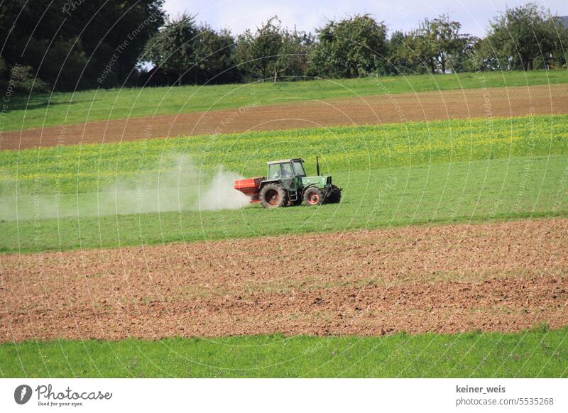 Ein Traktor fährt mit staubigem Dünger über das Feld und der Bauer betreibt die klassische Landwirtschaft Acker Staub Glyphosat Kalium grün Boden Erde Maschine