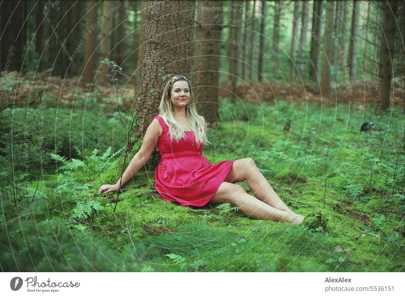 analoges Portrait einer schönen, blonden, langhaarigen Frau, die in einem lichten Wald an einem Baum gelehnt sitzt und in die Kamera schaut Gefühle attraktiv