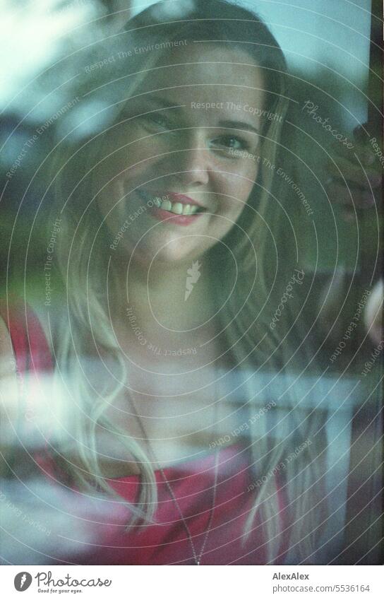 Blonde Frau schaut durch eine Fensterscheibe mit Reflexionen direkt in die Kamera und lächelt - analoges Portrait langhaarig lange Haare Reflexion & Spiegelung