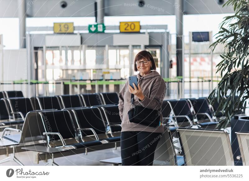 Glücklich lächelnde erwachsene Reisende Frau hält Smartphone, hat Videoanruf, winkt Hand zur Begrüßung. Aged Vlogger oder Blogger hat live am Flughafen-Terminal warten auf Flug, soziale Medien. Urlaub, Ferien