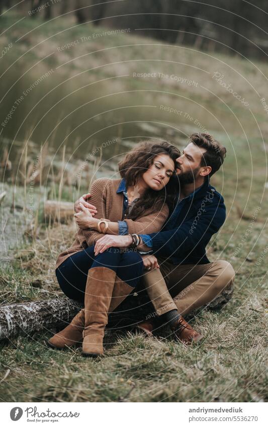 Glückliches Liebespaar sitzt am Ufer des Sees. Junges Paar umarmt sich an einem Herbsttag im Freien. Ein bärtiger Mann und eine lockige Frau in der Liebe. Valentinstag. Konzept der Liebe und Familie.