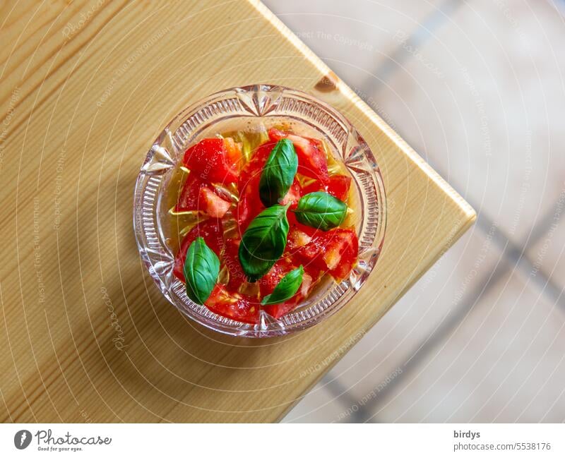 Frische Tomaten mit Olivenöl und frischem Basilikum in einer Glasschale Tomatensalat Basilikumblätter Tfrische Tomaten Tischkante Vogelperspektive Holztisch