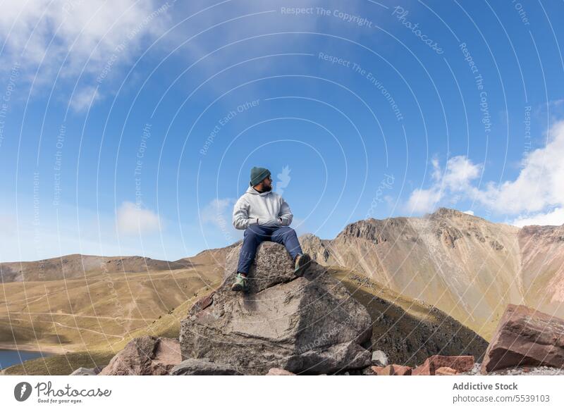 Junger Mann mit Mütze und warmer Kleidung sitzt auf einem Felsbrocken auf einem Berg Reisender Felsen Vulkan Landschaft Berge u. Gebirge bewundern erkunden