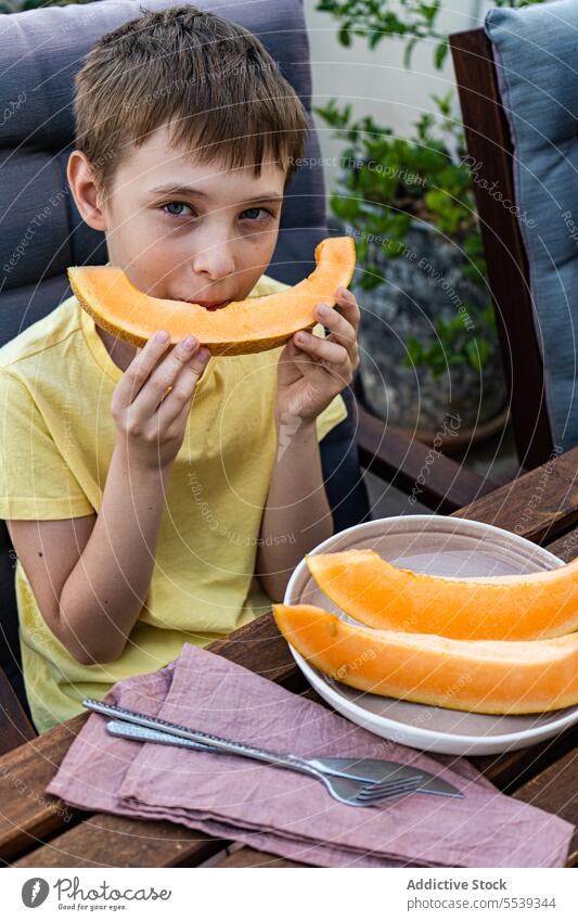 Preteen boy sitting at table and eating fresh cut musk melon Junge Kind essen Frucht frisch Gesundheit Melonen Scheibe Natur Sommer Eis Erfrischung trinken