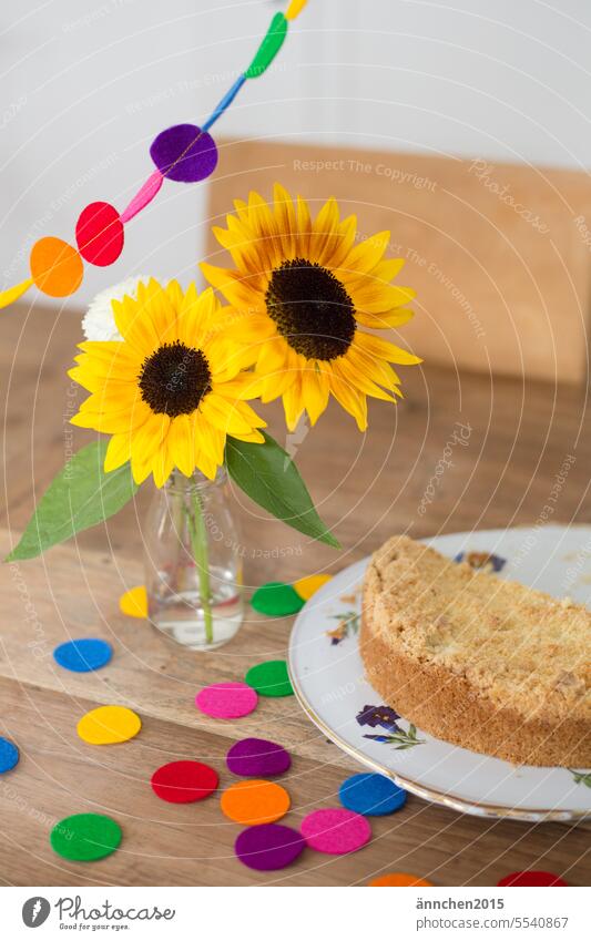 auf einem Holztisch stehen Sonnenblumen in einer kleinen Vase daneben steht ein Kuchen und man sieht Konfetti und eine bunte Girlande Strauß Blumen Feiern