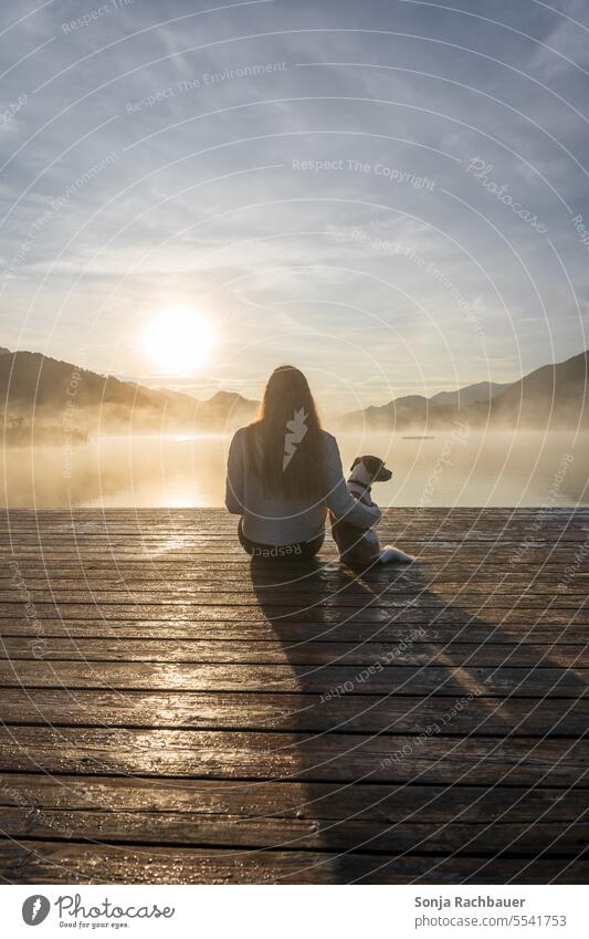 Eine junge Frau sitzt mit einem kleinen Hund auf einem Holzsteg an einem See. Sonnenaufgang, Rückansicht. sitzen Nebel Herbst Haustier Lifestyle Freundschaft
