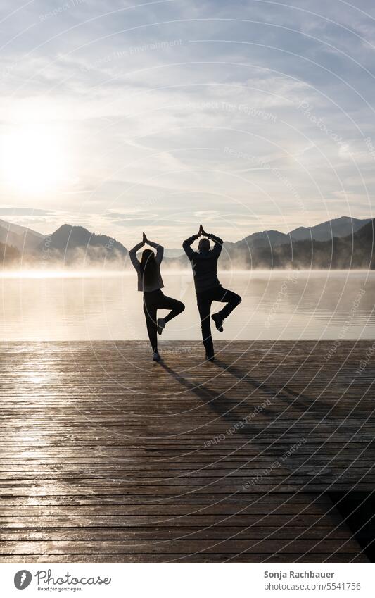 Vater und Tochter beim Yoga auf einem Holzsteg an einem See. Morgensonne, Herbst. Mann Frau Rückansicht Sonnenaufgang ruhig Idylle Nebel Morgenstimmung