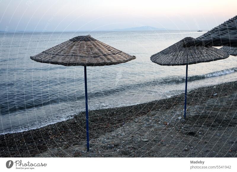 Blick vom Strand in Altinoluk auf Sonnenschirme aus Korbgeflecht im romantischen Licht der untergehenden Sonne und den Golf von Edremit am Ägäischen Meer in der Provinz Balikesir in der Türkei