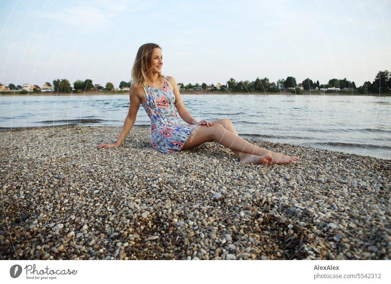Eine schöne, schlanke, junge Frau in einem Sommerkleid sitzt barfuß auf einer Sandbank aus Kies im Rhein und schaut in den Sonnenuntergang schöne Frau sportlich