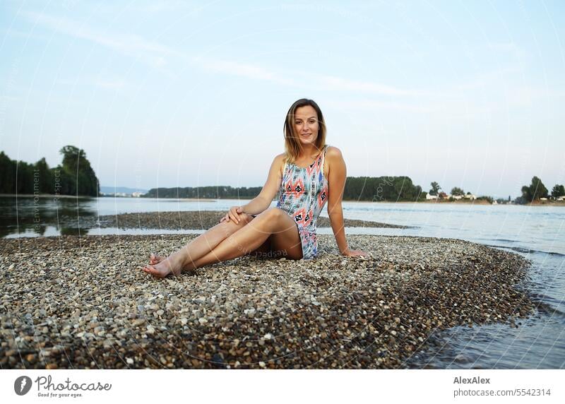 Eine schöne, schlanke, junge Frau in einem Sommerkleid sitzt barfuß auf einer Sandbank aus Kies im Rhein und schaut in den Sonnenuntergang schöne Frau sportlich