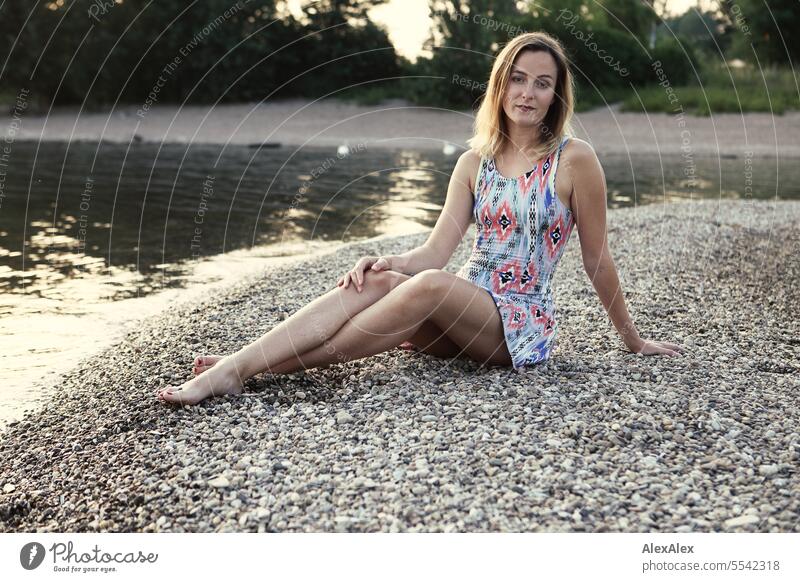 Eine schöne, schlanke, junge Frau in einem Sommerkleid sitzt barfuß auf einer Sandbank aus Kies im Rhein schöne Frau sportlich jugendlich 18-30 Jahre ästhetisch
