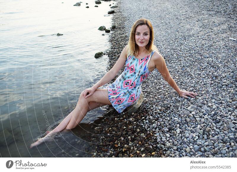 Eine schöne, schlanke, junge Frau in einem Sommerkleid sitzt barfuß am Rheinufer aus Kies mit den Füßen im Wasser schöne Frau sportlich jugendlich 18-30 Jahre