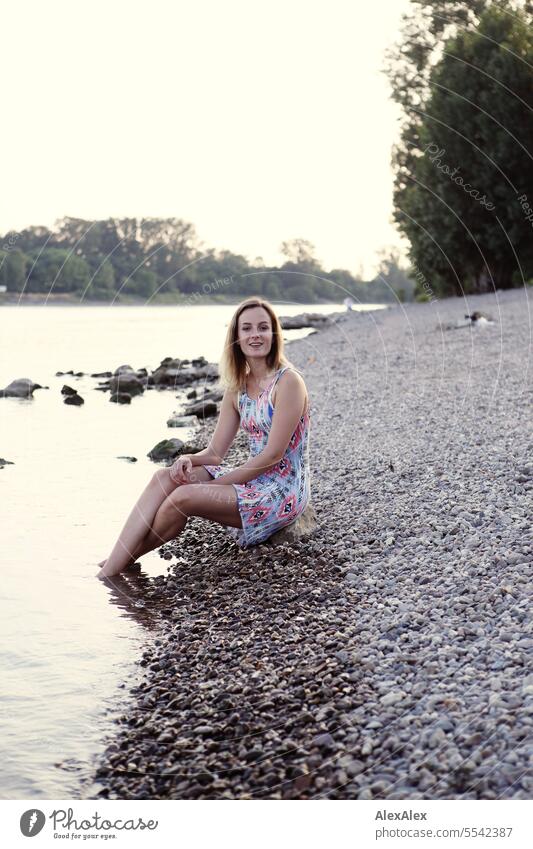 Eine schöne, schlanke, junge Frau in einem Sommerkleid sitzt barfuß am Rheinufer aus Kies mit den Füßen im Wasser schöne Frau sportlich jugendlich 18-30 Jahre