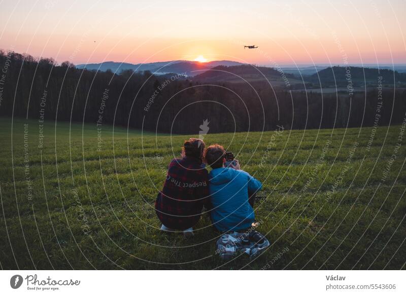 Ein Paar sitzt auf einer Anhöhe und genießt einen romantischen Abend. Ein Mann fliegt eine Drohne und nimmt einen atemberaubenden roten Sonnenuntergang auf