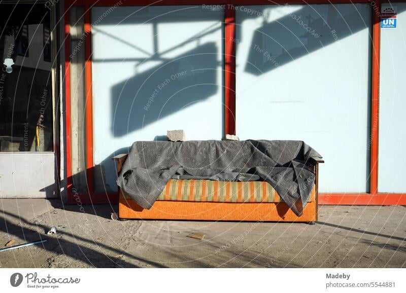 Altes ausrangiertes gestreiftes Sofa in Grün und Orange mit grauer Decke vor einem geschlossenen Geschäft im Sommer bei Sonnenschein in Adapazari in der Provinz Sakarya in der Türkei