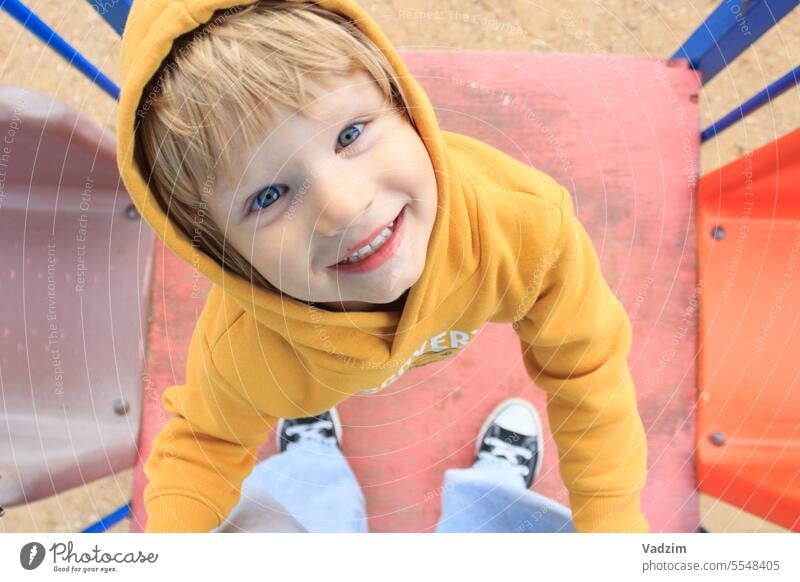 Ein fröhliches Kind in einem gelben Pullover spielt mit seiner Mutter auf dem Spielplatz, hält sich an ihren Beinen fest und schaut zu ihr auf. Familie Menschen