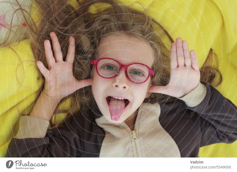 Ein fröhliches Mädchen mit Brille schneidet Grimassen, neckt sich und zeigt ihre Zunge mit ihren Händen in Form von Ohren, Großaufnahme. 8-10 Jahre alt