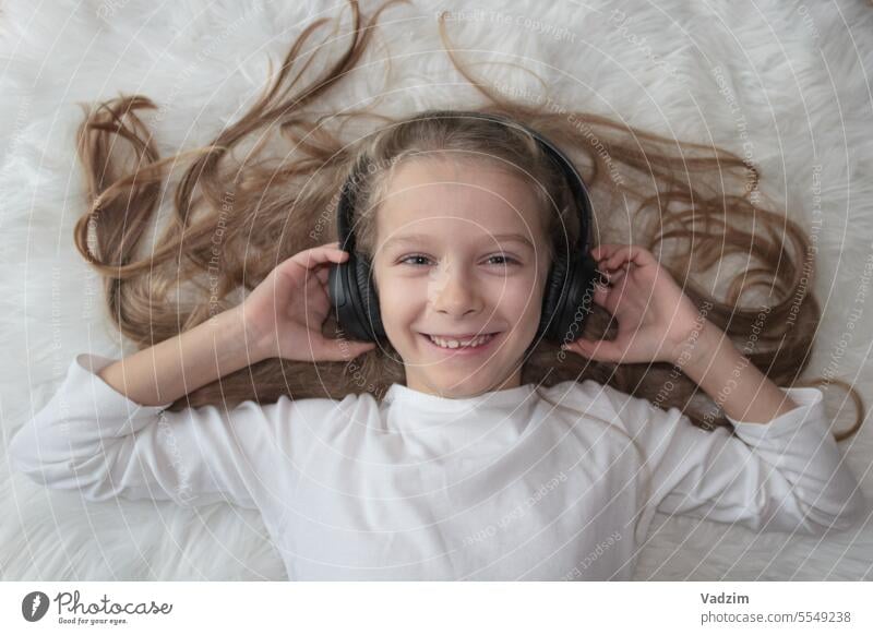 Kleines Mädchen mit kabellosen Kopfhörern auf einem weißen Fellteppich in Großaufnahme 5-10 Jahre alt Lifestyle Einführung der modernen Technologie Farbbild