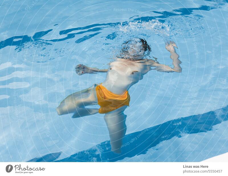 Ein einladender azurblauer Pool bildet die Kulisse für eine verspielte Szene. Ein barbusiges Mädchen befindet sich unter Wasser. Ihre sexy Kurven und ihr orangefarbener Bikini stehen im Mittelpunkt.