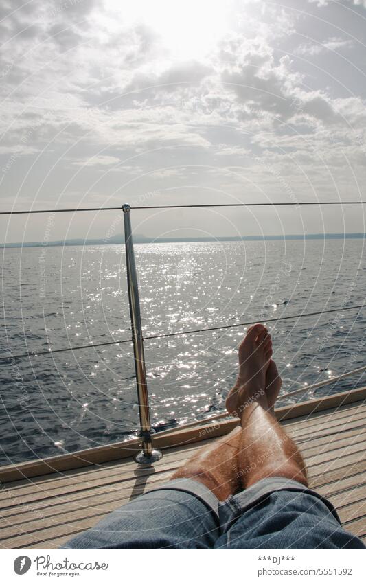 Mann mit kurzer Jeanshose auf einem Bootsdeck mit ausgestreckten, übereinander geschlagenen Beinen sitzend bz. liegend -  Blickrichtung hinaus aufs weite Meer.