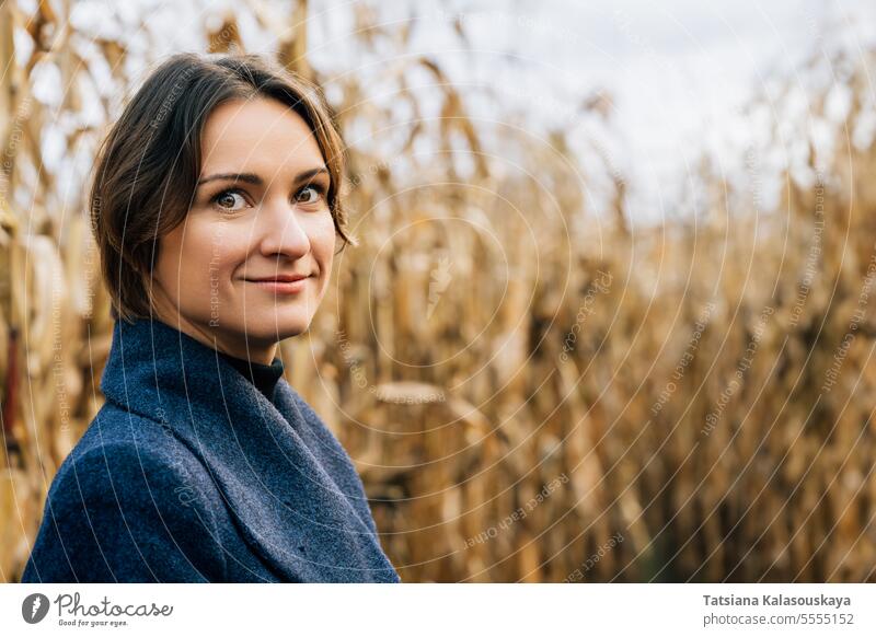 Porträt einer zuversichtlich lächelnden jungen Frau mit kurzen Haaren im Herbst inmitten eines Kornfelds selbstbewusst Lächeln kurzhaarig Mantel Reihen Mais