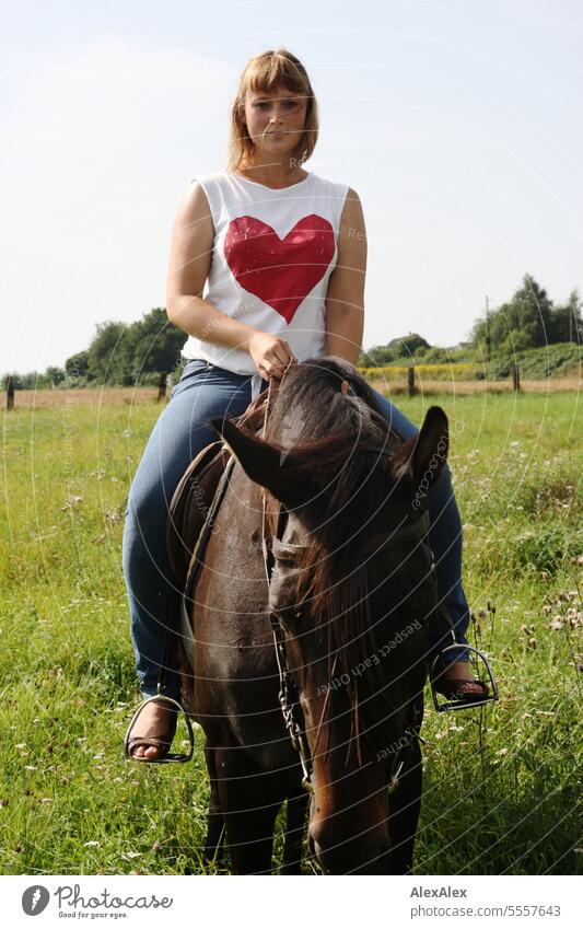 Portrait einer jungen Frau mit einem großen Herz auf dem T-Shirt, die auf einer grünen Weide auf einem braunen Pferd reitet junge Frau Kleid Garten schlank