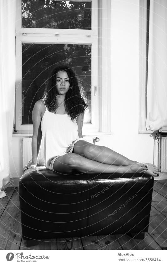 Eine junge, schöne Frau sitzt auf einem viereckigen Sofablock in einem Raum und schaut in die Kamera - schwarzweiß Aufnahme junge Frau langhaarig lockig Locken