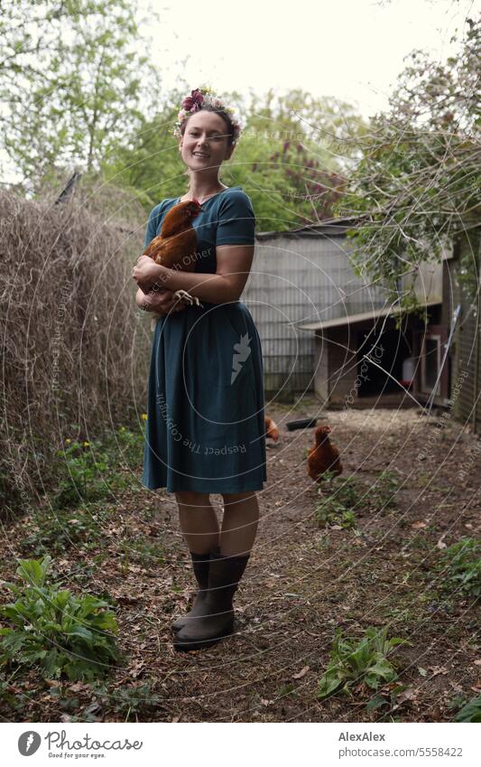 Portrait einer jungen Frau mit Blumenkranz im Garten mit einem braunen Huhn im Arm - in Gummistiefeln und Kleid im Hühnerauslauf junge Frau Beine schlank