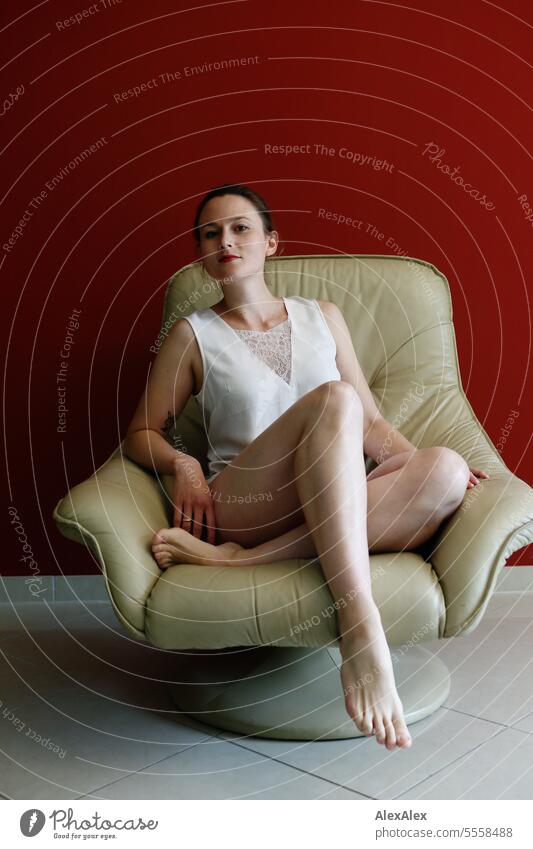 Junge, schlanke, sportliche Frau sitzt in einem beigen Ledersessel vor einer roten Wand und posiert für die Kamera junge Frau schön fit Beine muskulös Haut